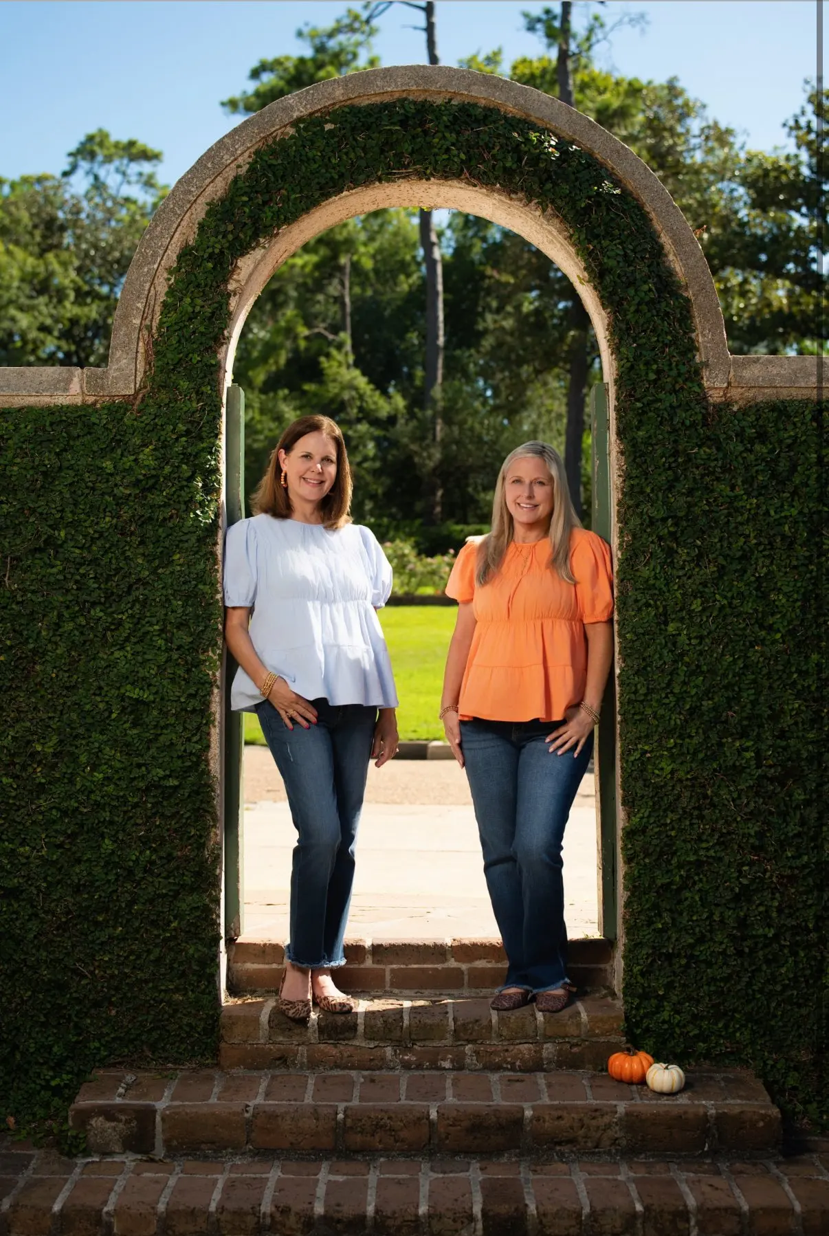 Michelle and Stephanie standing in an ivy covered garden arch with two small pumpkins at their feet