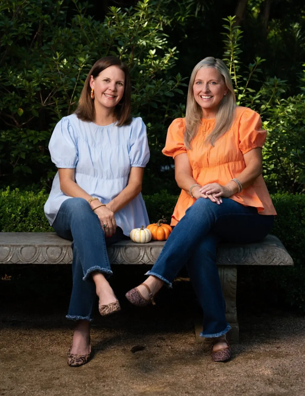 Michelle and Stephanie seated on a stone garden bench with a white and an orange pumpkin between them